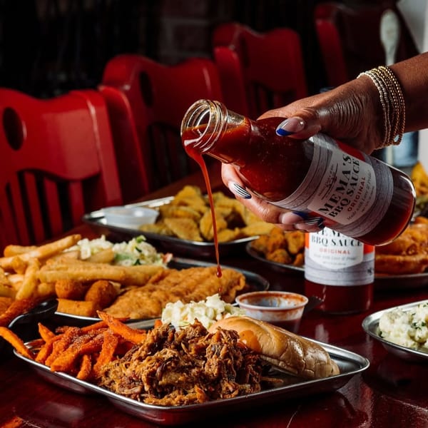 Hand pours Ken’s Original barbecue sauce over platters of fried seafood, pulled pork sandwich, sweet potato fries, and coleslaw.