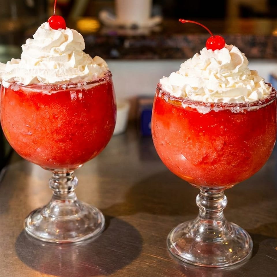 Two red frozen drinks in stemmed glasses topped with whipped cream and cherries on a restaurant counter.