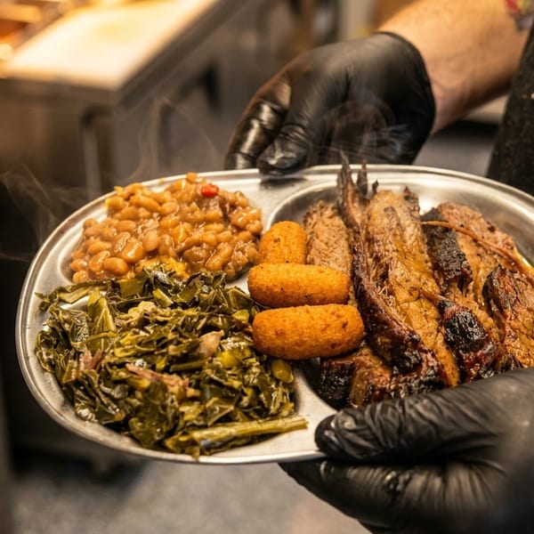 Gloved server presents a steaming platter of grilled meats, baked beans, collard greens, and golden cornbread in a restaurant kitchen.