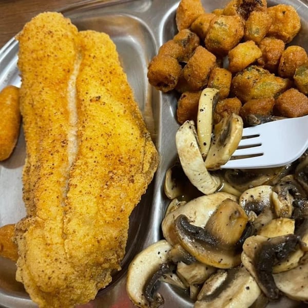 Breaded fish fillets with fried vegetables and sautéed mushrooms served in a compartmented metal tray.