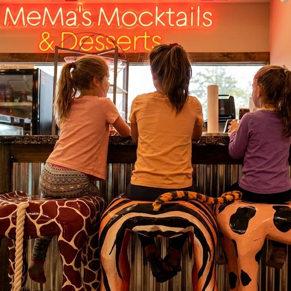 Three children sit on animal-themed stools at a dessert counter beneath a neon sign advertising mocktails and desserts.