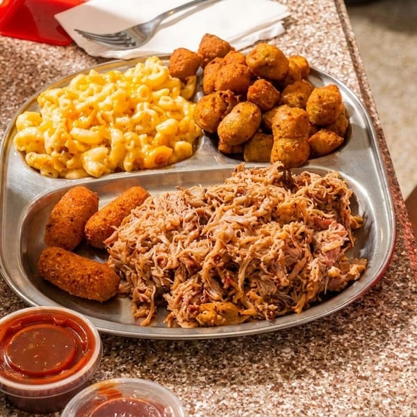 Diner plate featuring macaroni and cheese, fried bites, croquettes, and shredded meat with dipping sauces and iced soda.