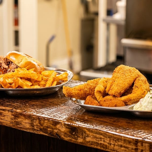 Two metal plates of comfort food featuring pulled pork sandwiches, fries, fried seafood, hush puppies, and coleslaw on a rustic restaurant counter.