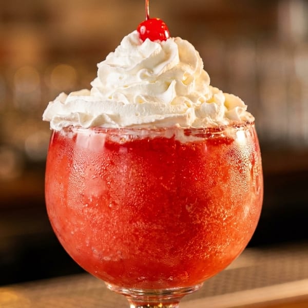 Red frozen cocktail topped with whipped cream and a cherry, served in a rounded glass at a warmly lit bar.