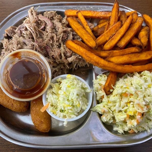 Tray of pulled pork, sweet potato fries, hush puppies, coleslaw, and dipping sauce on a wooden table.