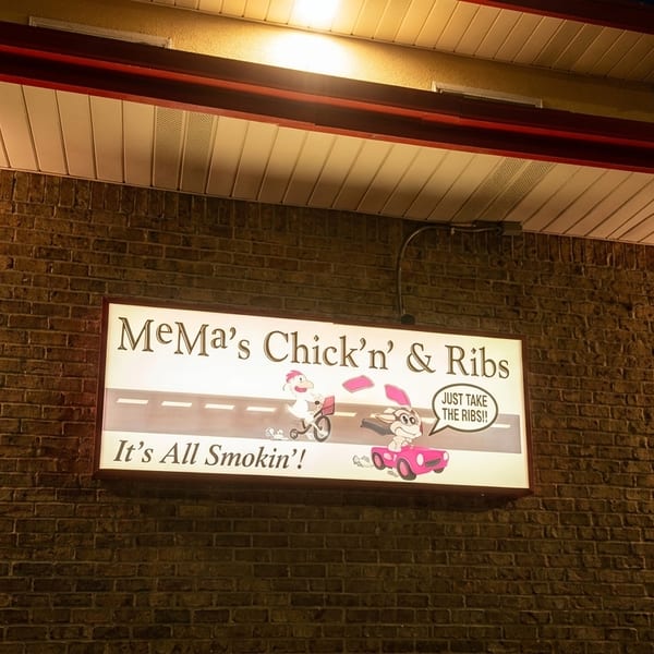 Illuminated MeMa’s Chick’n’ Ribs restaurant sign mounted on a brick wall at night, featuring cartoon characters and a smoking barbecue slogan.