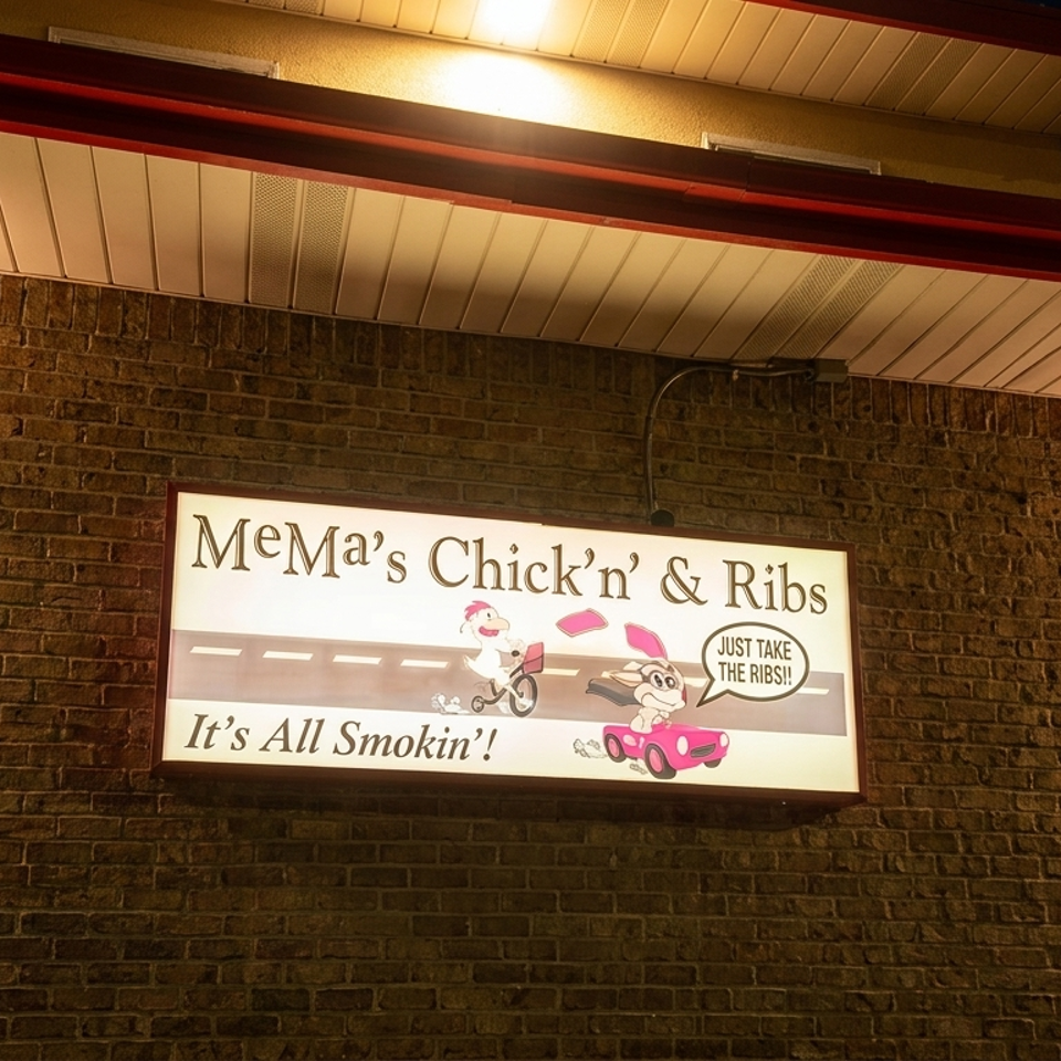 Illuminated MeMa’s Chick’n’ Ribs restaurant sign mounted on a brick wall at night, featuring cartoon characters and a smoking barbecue slogan.
