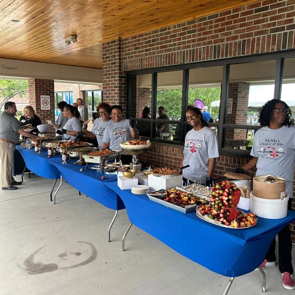 People serve barbecue, sides, and colorful fruit at a MeMa’s Chick’n & Ribs buffet beneath a covered brick patio.