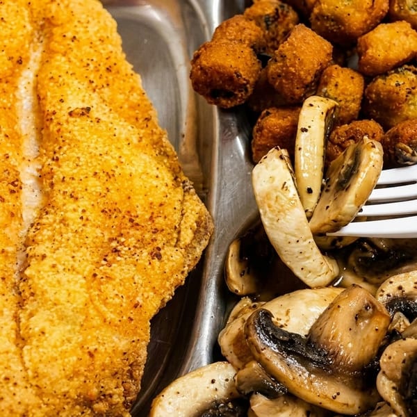 Crispy breaded fish fillets served with fried hushpuppies and sautéed mushrooms on a divided metal tray.